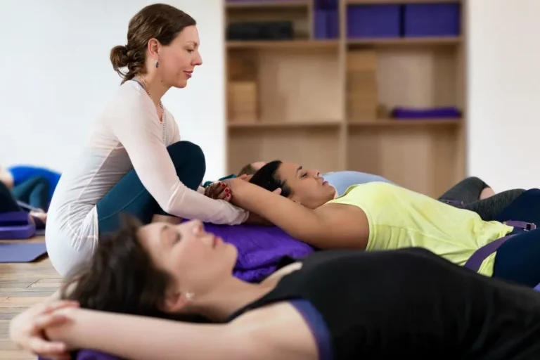 A group of people are practicing restorative yoga in a Triyoga studio in London. They are lying on yoga mats, supported by bolsters, blankets, and blocks. Ann, the yoga therapist, is leading the class. She is correcting the posture f a student, giving instructions. The students are all relaxed and focused. The atmosphere is peaceful and serene.