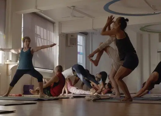 several people and an instructor are practicing Mysore style Ashtanga yoga in a yoga studio in London. Each person is doing a different pose, and the instructor is guiding them.