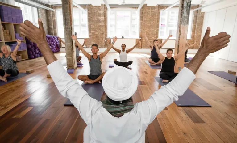 A yoga teacher is leading a yoga class in Triyoga studio, London. The teacher is standing in front of a group of students who are all practicing yoga poses. The students are all wearing comfortable clothing and have focused expressions on their faces. The yoga class is taking place in a large, bright room with two columns and large windows letting in a lot of natural sunlight.