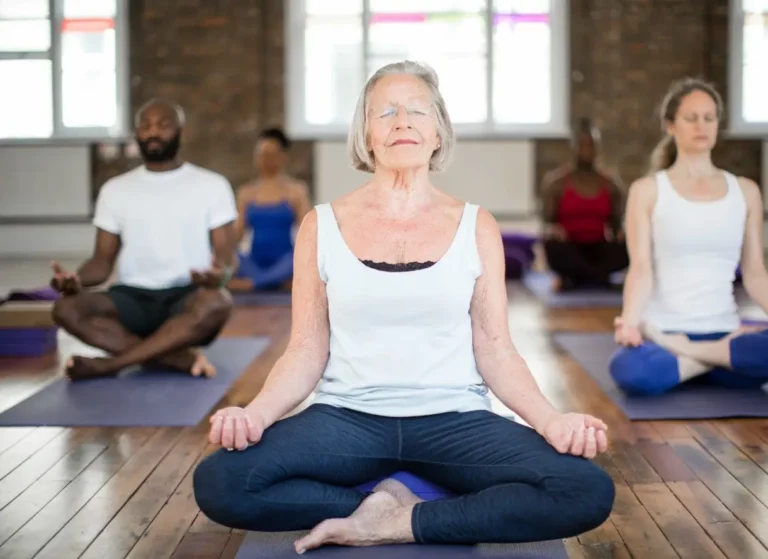 An elderly woman leads a group of students in a yoga and meditation class at Triyoga London. The students are all different ages and abilities, and they are all focused on their practice. The woman is smiling and encouraging, and she is clearly passionate about teaching yoga. The students are relaxed and peaceful, and they are clearly benefiting from the class.