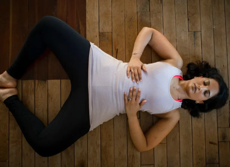 A woman in white clothing is laying back on a wooden floor at Triyoga. She is practicing yin yoga, a type of yoga that focuses on holding postures for longer periods of time. Her eyes are closed and she appears to be relaxed and at peace.