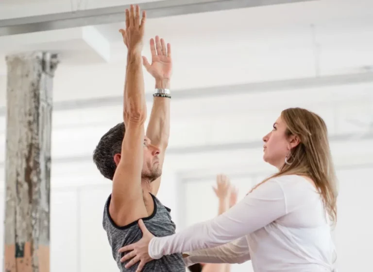 A new yoga student is being assisted by a yoga instructor. The student is standing with his hands raised, and the instructor is helping him with his posture and breathing. The student looks focused and determined, and the instructor looks supportive and encouraging.